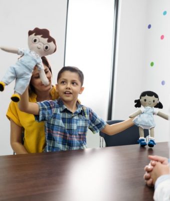 A little latin boy sitting on his mother's lap and holding learning dolls at the psychologist consultation office.