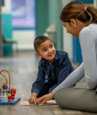 A young mixed race boy looks at his speech therapist while pointing at a book in front of him.